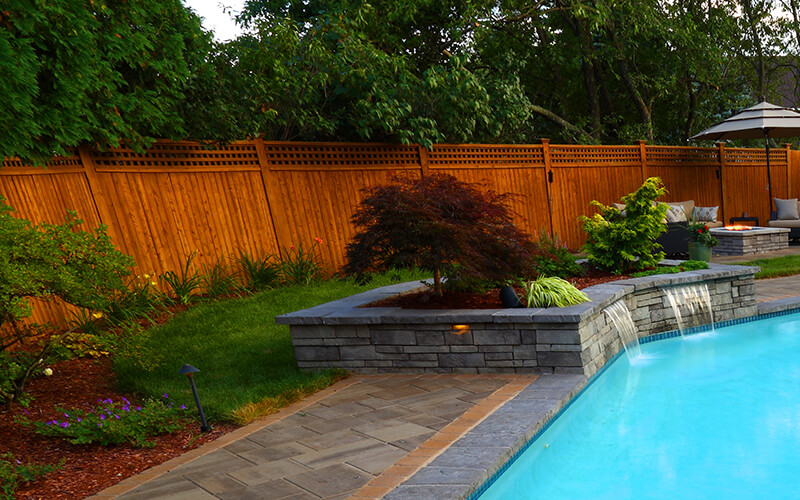 This image depicts a luxurious backyard setup that combines a pool, seating area, and fire pit to create an ideal space for outdoor relaxation. In the foreground, there is a square fire pit made of stacked stone with a smooth concrete top. The fire pit is lit, with flames dancing over blue glass stones, adding a vibrant and cozy ambiance to the scene. Surrounding the fire pit are comfortable wicker chairs with beige cushions, offering seating for relaxation and warmth. The patio area is paved with large stone tiles in warm tones, seamlessly blending with the natural elements of the space. In the background, a pristine blue swimming pool stretches out, with several white lounge chairs and umbrellas positioned on a deck area near the pool's edge. A grassy section adds greenery between the pool and the patio, while planters with vibrant plants and shrubs contribute to the lush, landscaped look. The entire space is framed by a backdrop of trees and hedges, providing a sense of privacy and seclusion. This setting combines comfort and elegance, perfect for outdoor gatherings or quiet evenings by the fire. This scene has been carefully crafted by Landscape architects Jim and Jennifer DiNovo of Design Works Landscaping in Hudson New Hampshire. Design Works offers landscaping to homeowners living in Southern New Hampshire, and has been offering their services for nearly two decades.